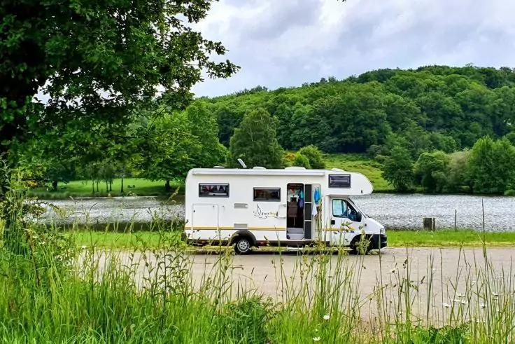 Wei&szlig;es Wohnmobil mit ge&ouml;ffneter T&uuml;r auf Parkfl&auml;che am Ufer eines Sees, im Vordergrund Gras, im Hintergrund B&auml;ume und bewaldeter H&uuml;gel unter wolkigem Himmel