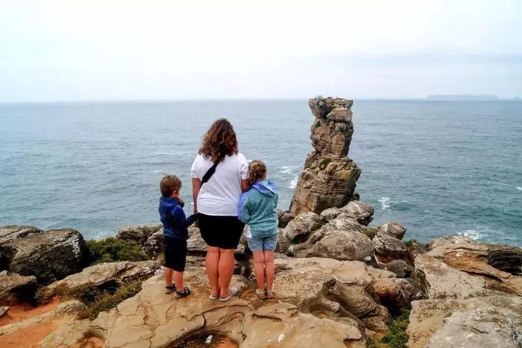 Drei Personen (eine Erwachsene, zwei Kinder) stehen auf einer felsigen Klippe und blicken aufs Meer. Erwachsene: wei&szlig;es Oberteil, schwarze Shorts; Kinder: blaues bzw. hellblaues Oberteil; Felsen im Vordergrund