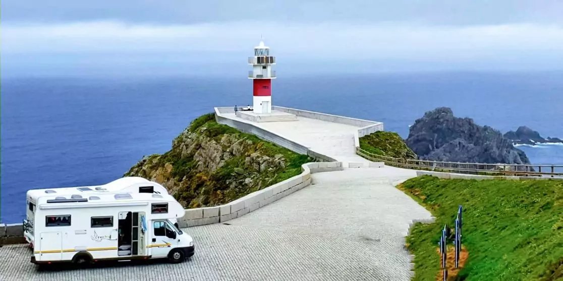 Wei&szlig;es Wohnmobil auf gepflastertem Platz neben einem Leuchtturm mit rotem Streifen auf einem Felsvorsprung, Meer, Himmel und grasbewachsene Hang sichtbar