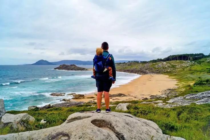 Person mit Kind in R&uuml;ckentrage steht auf einem Felsen; im Vordergrund Felsen, im Hintergrund Sandstrand, Meer mit Wellen, bew&ouml;lkter Himmel und entfernte H&uuml;gel