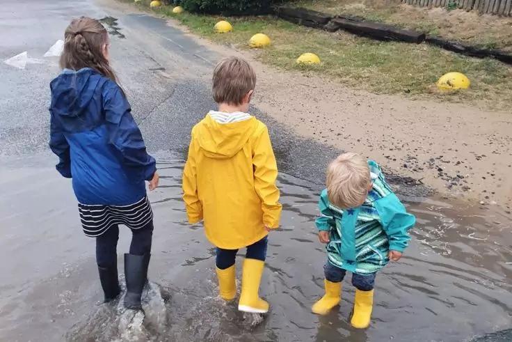 Drei Kinder von hinten in Regenm&auml;nteln und Gummistiefeln stehen in einer Pf&uuml;tze auf einer nassen Stra&szlig;e
