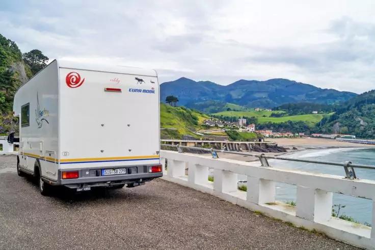 Wei&szlig;es Wohnmobil steht an einer K&uuml;stenpromenade, im Hintergrund Meer, Pier, H&auml;user und gr&uuml;ne Berge unter wolkigem Himmel