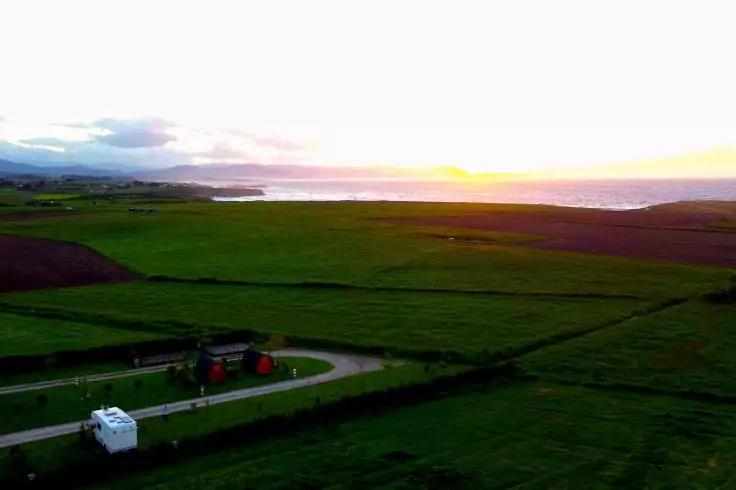 Luftaufnahme gr&uuml;ner Felder mit geschwungener Stra&szlig;e, wei&szlig;em Wohnwagen, zwei roten Geb&auml;uden, K&uuml;ste und Sonne am Horizont