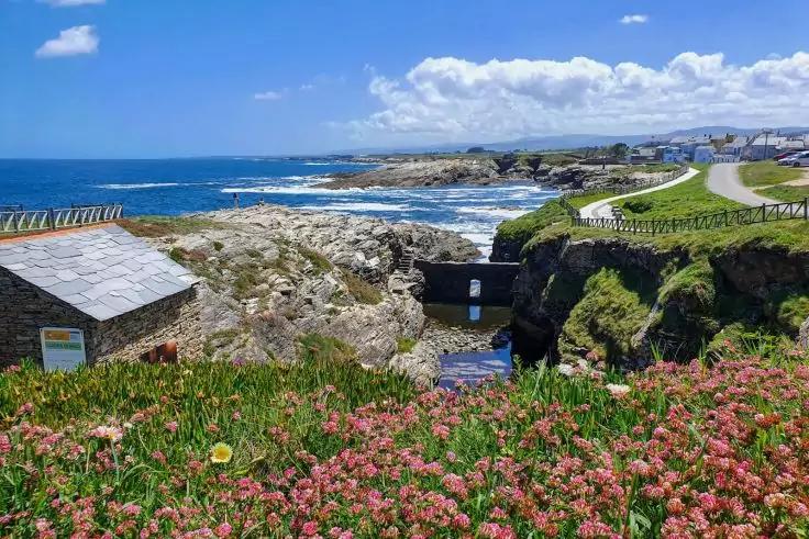 Felsige K&uuml;ste mit rosa Bl&uuml;ten im Vordergrund, kleines steinernes Geb&auml;ude links, schmaler Wasserkanal zur Brandung, Gr&uuml;nfl&auml;chen und K&uuml;stenweg mit Pfad, blaues Meer und teils bew&ouml;lkter Himmel, entfernte H&auml;user am Horizont