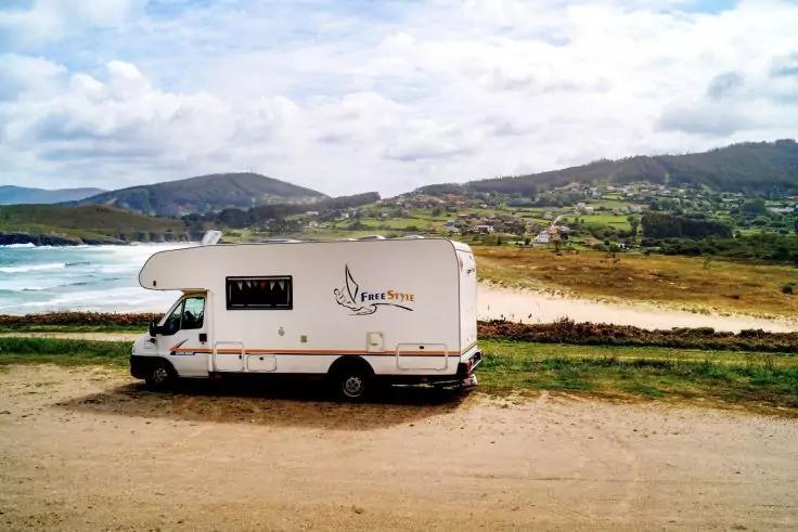 Wei&szlig;es Reisemobil auf unbefestigtem Platz am Meer, Sandstrand, Wellen, H&uuml;gel mit H&auml;usern und bew&ouml;lktem Himmel