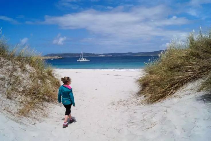 Eine Person in einer t&uuml;rkisfarbenen Jacke mit rosa Kapuze und schwarzen Shorts geht &uuml;ber einen sandigen Pfad zwischen grasbewachsenen D&uuml;nen zum wei&szlig;en Sandstrand; blaues Meer mit einem Segelboot in der Ferne und teilweise bew&ouml;lkter Himmel