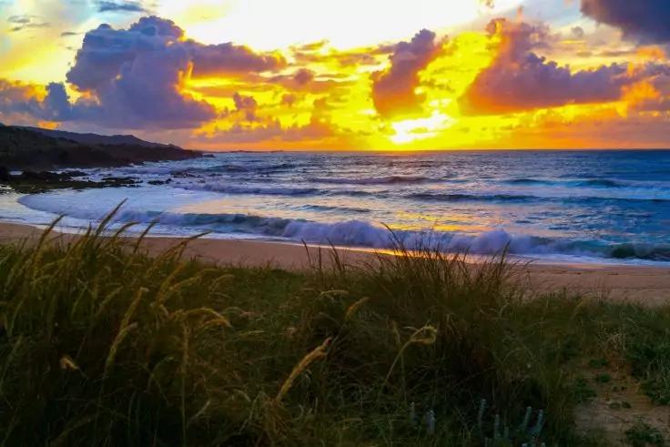 Sandstrand mit D&uuml;nengras im Vordergrund, Wellen, Felsen links und die Sonne knapp &uuml;ber dem Meereshorizont mit gelb-orangefarbenem Himmel und Wolken
