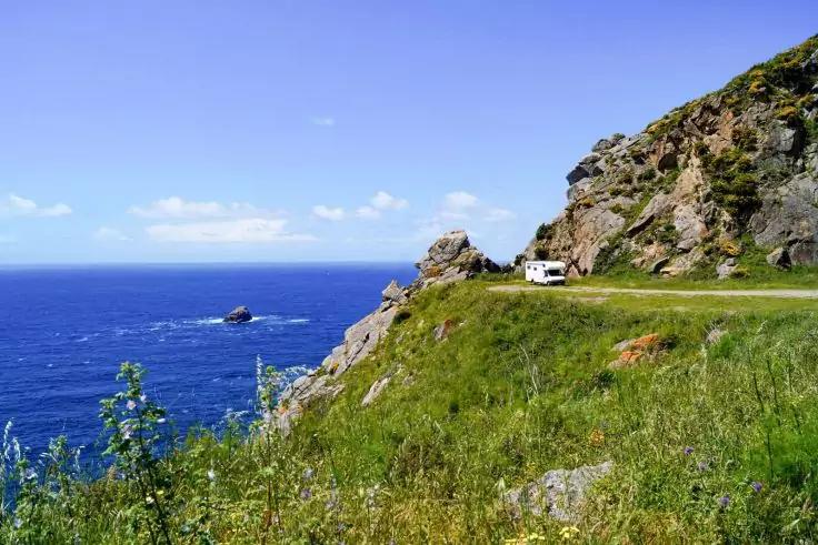 K&uuml;stenstra&szlig;e auf Klippe mit grasbewachsenem Abhang, Felsformationen, offenes Meer mit kleinem Felsen im Wasser, wei&szlig;es Wohnmobil auf der Stra&szlig;e, Himmel mit wenigen Wolken