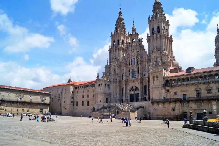 Gro&szlig;e steinerne Fassade mit zwei hohen T&uuml;rmen, Treppen zum Haupteingang, weiter gepflasterter Platz mit vereinzelten Personen und blauem Himmel mit Wolken