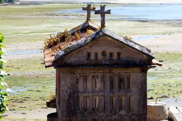 Kleiner rechteckiger steinerner Bau mit Satteldach und zwei Kreuzen auf dem Dach, Moos und Pflanzen auf dem First, Vorderseite mit vertikalen Platten, im Hintergrund Watt und flaches Wasser