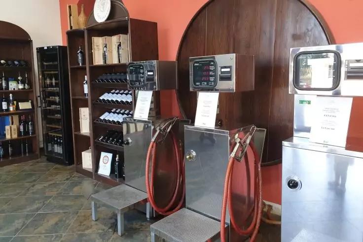 Innenraum mit zwei Edelstahlger&auml;ten mit roten Schl&auml;uchen und digitalen Anzeigen auf Podesten, rechts ein weiteres Edelstahlger&auml;t, links Regale mit Glasflaschen und Kartons, runde Holzverkleidung an der Wand, Fliesenboden