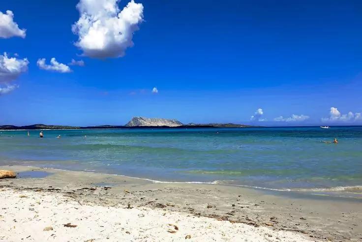 Sandstrand im Vordergrund, flaches t&uuml;rkisfarbenes Meer, kleine Insel am Horizont, blauer Himmel mit wenigen wei&szlig;en Wolken