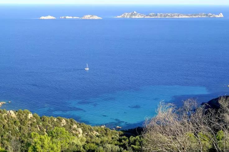 K&uuml;ste mit tiefblauem Meer, ein Segelboot in der Ferne, kleine Inseln am Horizont und bewachsene Felsen im Vordergrund