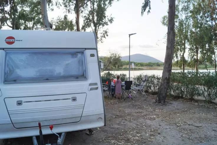 Wohnwagen links; drei Personen auf Campingstühlen unter Bäumen am Seeufer; Hügel im Hintergrund