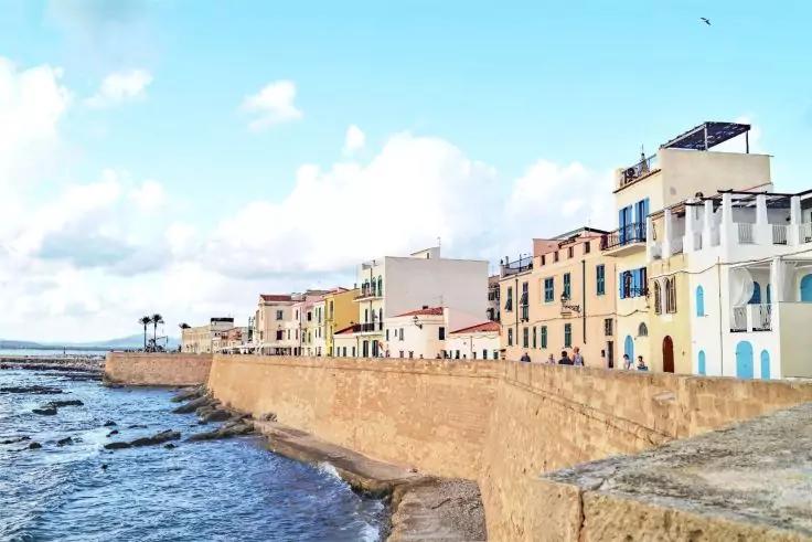 Reihe farbiger mehrstöckiger Häuser entlang einer steinernen Ufermauer, Meer mit Wellen links, bewölkter Himmel, einige Personen auf der Mauer, Vogel am Himmel