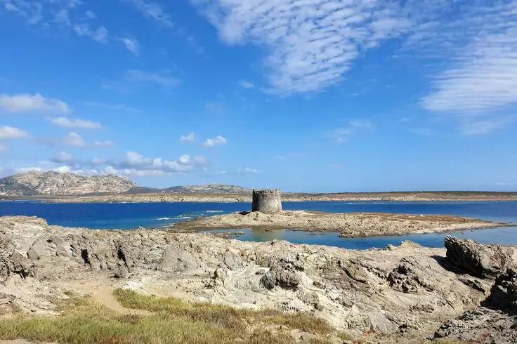 Küstenlandschaft mit Felsen im Vordergrund, rundem Steinturm auf einer kleinen Insel und blauem Himmel