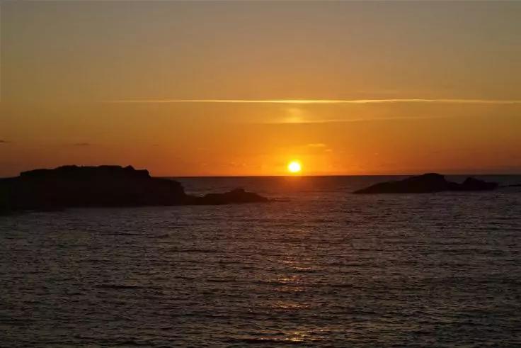 Sonnenuntergang über dem Meer, Sonne knapp über dem Horizont, dunkle Felsen am Horizont, wellige Wasseroberfläche