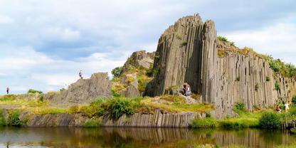 S&auml;ulenartige Felsformation am Ufer mit gr&uuml;ner Vegetation, Wasser mit Spiegelung, vereinzelte Personen auf den Felsen und bew&ouml;lkter Himmel