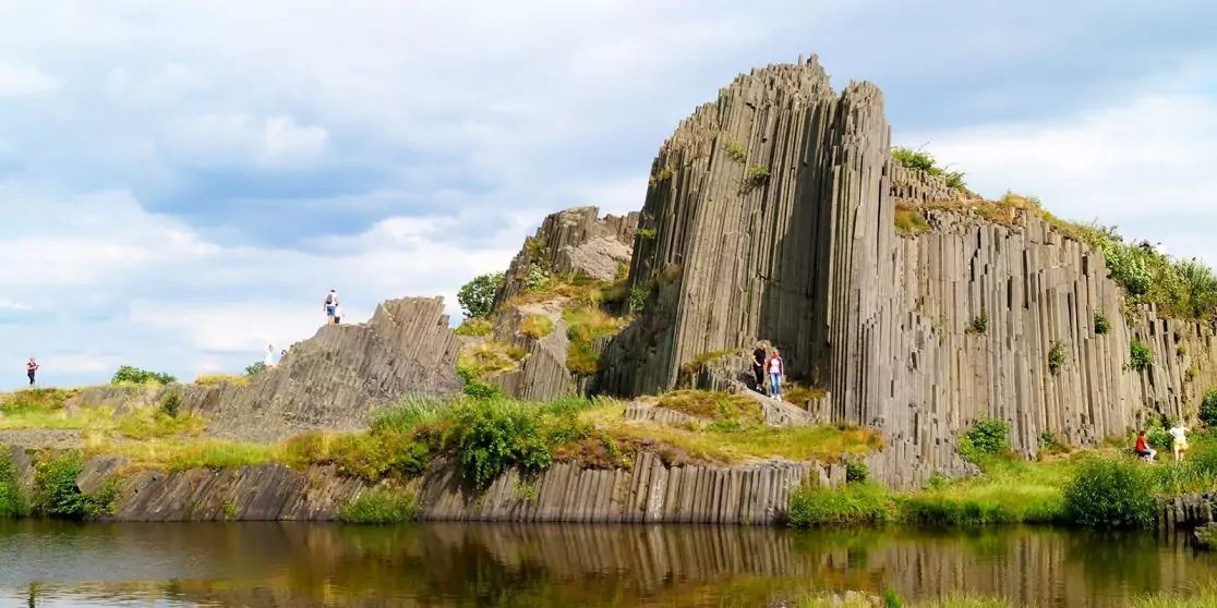 S&auml;ulenartige Felsformation am Ufer mit gr&uuml;ner Vegetation, Wasser mit Spiegelung, vereinzelte Personen auf den Felsen und bew&ouml;lkter Himmel