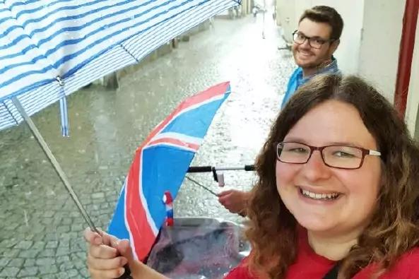 Zwei Personen mit Regenschirmen auf nassem Kopfsteinpflaster; vorn eine l&auml;chelnde Person mit Brille und rotem Oberteil, hinten eine Person mit Brille und blauem Oberteil. Ein Regenschirm zeigt die britische Flagge, oben ein blau-wei&szlig; gestreifter Mark