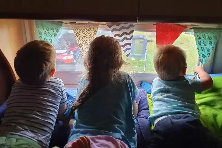 Drei Kinder von hinten sitzen nebeneinander vor einem Fenster, bunte Wimpel dar&uuml;ber, Autos und Rasen drau&szlig;en sichtbar