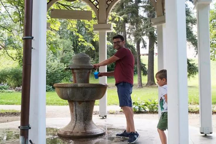 Mann f&uuml;llt eine Flasche an einem steinernen Trinkbrunnen in einem offenen Pavillon, ein Kind steht daneben