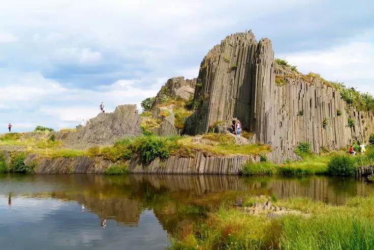 S&auml;ulenf&ouml;rmige Felsformation am Ufer, ruhiges Wasser mit Spiegelung, grasbewachsene Uferbereiche, mehrere Personen auf und neben den Felsen, bew&ouml;lkter Himmel