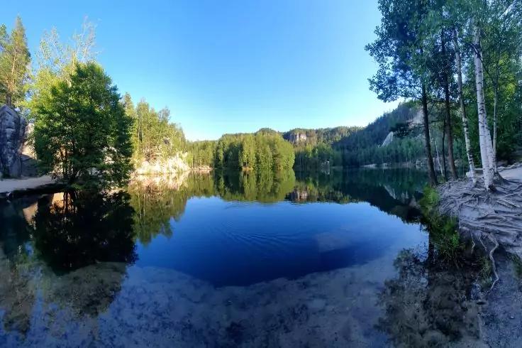 See mit Spiegelung von B&auml;umen, umgebenden Felsen, blauem Himmel, Birken am rechten Ufer und flachem, steinigem Vordergrund