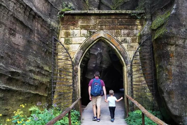 Zwei Personen, eine mit blauem Rucksack und eine kleinere an der Hand, gehen &uuml;ber eine Holzbr&uuml;cke durch einen steinernen spitzbogigen Torbogen in eine dunkle Fels&ouml;ffnung; Felsw&auml;nde und Pflanzen sichtbar