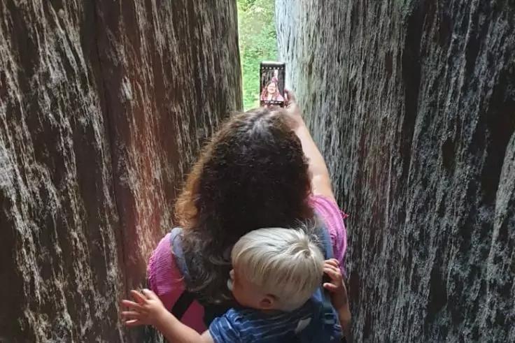 Erwachsene Person mit lockigem Haar tr&auml;gt ein blondes Kleinkind in einer Trage und h&auml;lt ein Smartphone hoch zwischen zwei hohen Felsw&auml;nden