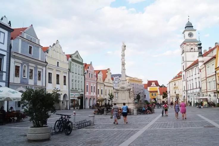 Platz mit Kopfsteinpflaster, Reihe pastellfarbener Giebelh&auml;user, zentrale S&auml;ule mit Figuren, Uhrturm rechts, Menschen zu Fu&szlig;, geparkte Fahrr&auml;der und Au&szlig;ensitzpl&auml;tze