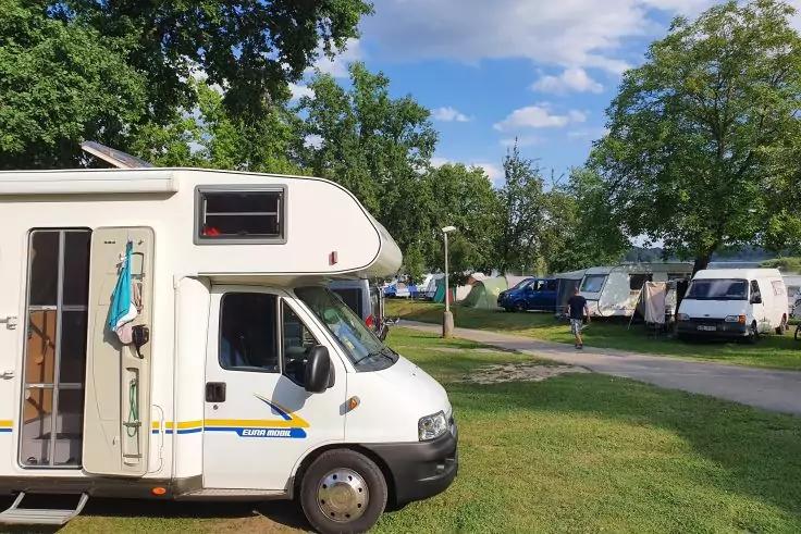 Wei&szlig;es Wohnmobil mit offener T&uuml;r und aufgeh&auml;ngtem Handtuch auf einem grasbewachsenen Campingplatz, weitere Wohnwagen, B&auml;ume und blauer Himmel mit Wolken im Hintergrund
