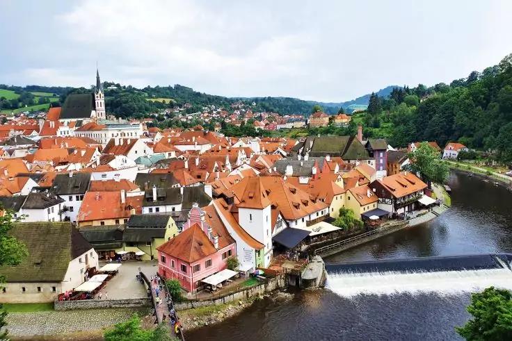 Ufer einer historischen Stadt mit eng stehenden roten Ziegeld&auml;chern, einer Kirche mit Turm, einem Fluss mit kleinem Wehr/dam und gr&uuml;nen H&uuml;geln im Hintergrund