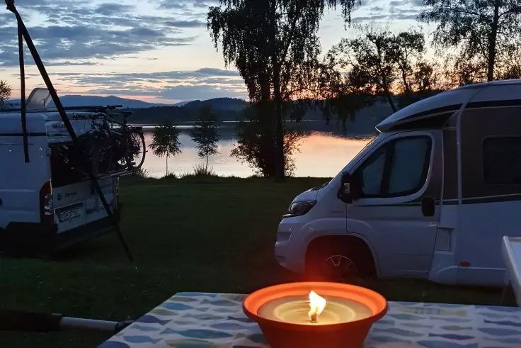 Brennende Kerze in einer Schale auf einem Tischtuch im Vordergrund; im Hintergrund zwei Wohnmobile am Ufer eines Sees, B&auml;ume, H&uuml;gel, wolkiger Himmel mit r&ouml;tlich-blauen Farbt&ouml;nen und Spiegelung im Wasser