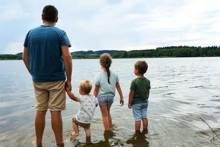 Ein Mann steht mit drei Kindern knietief in einem See, R&uuml;cken zur Kamera; Ufer und bew&ouml;lkter Himmel im Hintergrund