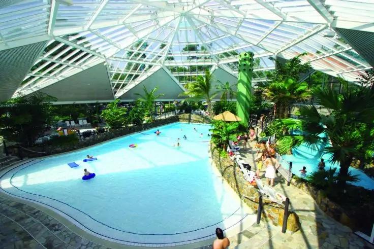 &Uuml;berdachtes Schwimmbad unter gl&auml;serner Kuppel, Palmen, mehrere Personen im Wasser und Liegen am Beckenrand