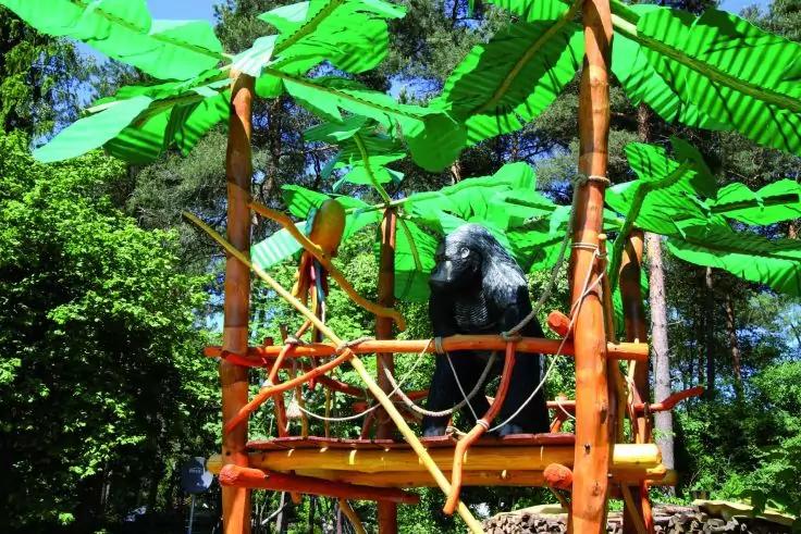 Schwarze Gorillafigur auf h&ouml;lzernem Spielpodest mit gelben und orangen Elementen, gro&szlig;e gr&uuml;ne Bl&auml;tter dar&uuml;ber