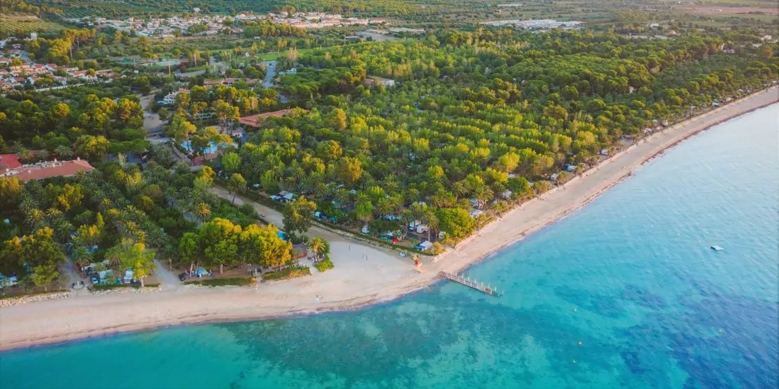 Luftaufnahme einer sandigen K&uuml;ste mit t&uuml;rkisfarbenem Meer, Strand, Holzsteg und von B&auml;umen umgebenen H&auml;usern