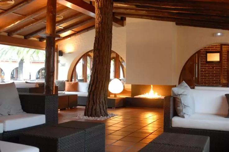 Innenraum mit Sofas, Kamin, sichtbarem Baumstamm, Holzdeckenbalken, Fliesenboden und runder Lampe