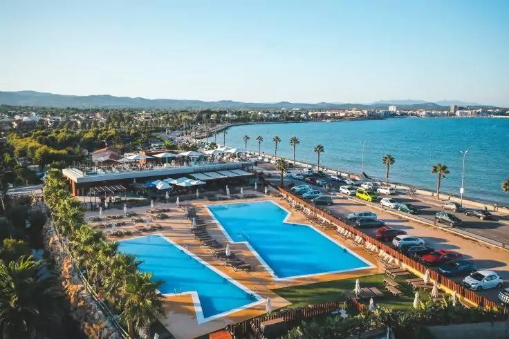 Freiluft-Schwimmbad mit zwei rechteckigen Becken, Liegen und Palmen neben einer Stra&szlig;e mit geparkten Autos, Meer und K&uuml;stenlinie im Hintergrund