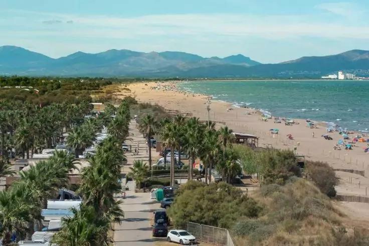 Promenade mit Palmen und geparkten Autos neben einem sandigen Strand mit Sonnenschirmen, Menschen, D&uuml;nen, Meer und Bergen im Hintergrund