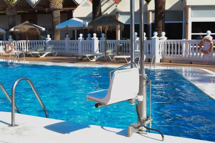 Au&szlig;enpool mit blauem Wasser, Metall-Einstiegsleiter, Poollift mit Sitz am Beckenrand, Sonnenliegen, Sonnenschirme und Strohd&auml;cher, wei&szlig;e Balustrade, Geb&auml;ude und Palmen im Hintergrund, Rettungsring an einer S&auml;ule