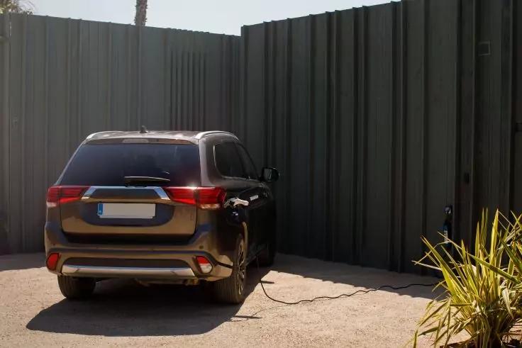 Dunkles SUV von hinten links geparkt neben einem hohen grauen Metallzaun, ein Kabel verl&auml;uft vom Heck zu einer kleinen schwarzen Box am Zaun, Pflanzen im rechten Vordergrund