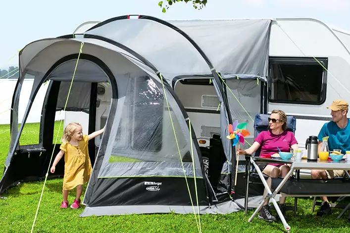 Wohnwagen mit angebautem Vorzelt auf einer Wiese am Wasser; ein kleines Mädchen steht am Eingang, zwei Erwachsene sitzen an einem Campingtisch mit Bechern und Picknick