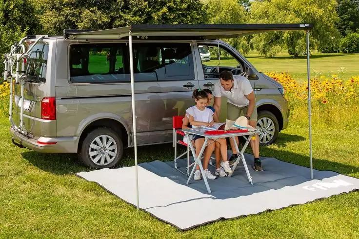 Grauer Camper-Van mit ausgefahrener Markise auf einer Wiese; auf einer Bodenmatte davor zwei Personen (Erwachsener und Kind) an einem Campingtisch mit Laptop und Buch; im Hintergrund Bäume, gelbe Blumen und ein Fahrradträger am Heck