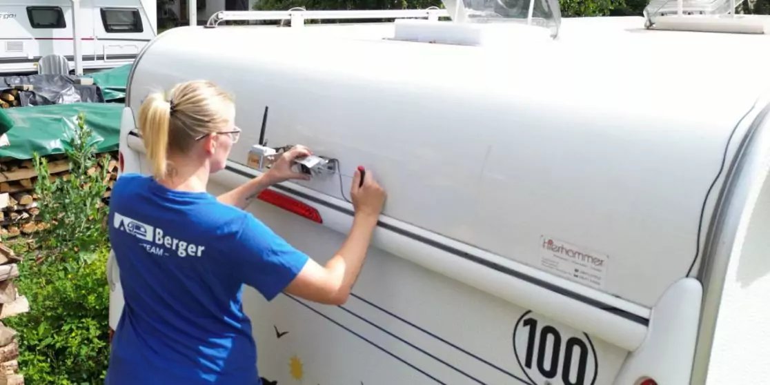 Person mit blondem Zopf und blauem T‑Shirt steht an der Seite eines wei&szlig;en Wohnwagens und h&auml;lt mit beiden H&auml;nden eine seitliche Klappe