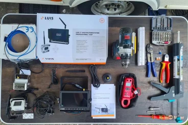 Tisch mit Elektronik- und Handwerkzeugen: Karton mit Aufschrift "LUIS", Kabelrollen, kleines Display, rotes Messgerät, Elektrowerkzeug, Bit-Set, Zangen, Schraubendreher, Heißklebepistole
