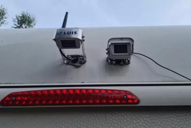 Zwei rechteckige Kameragehäuse an der weißen oberen Fahrzeugkante, linke Kamera mit Aufschrift 'LUIS', schwarzes Kabel an der rechten Kamera, darunter rotes LED-Lichtband, Himmel mit Baumkronen