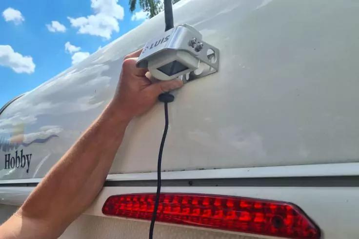 Eine Hand hält ein silbernes rechteckiges Gerät mit schwarzem Kabel an der Außenwand eines weißen Wohnwagens; darunter ein rotes Rücklicht und blauer Himmel mit Wolken
