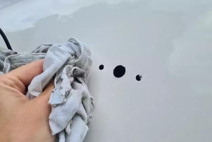 Hand hält ein helles Tuch auf einer glatten weißen Oberfläche neben drei runden schwarzen Flecken